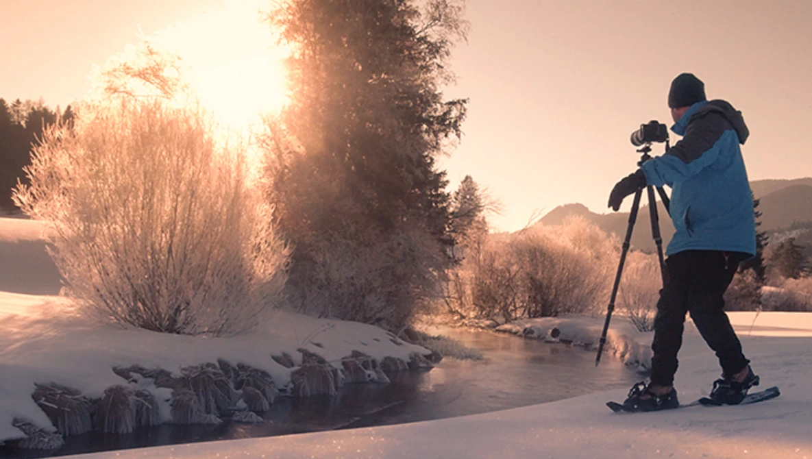 Photographer on skis operating a camera on tripod in snowy landscape at sunrise.
