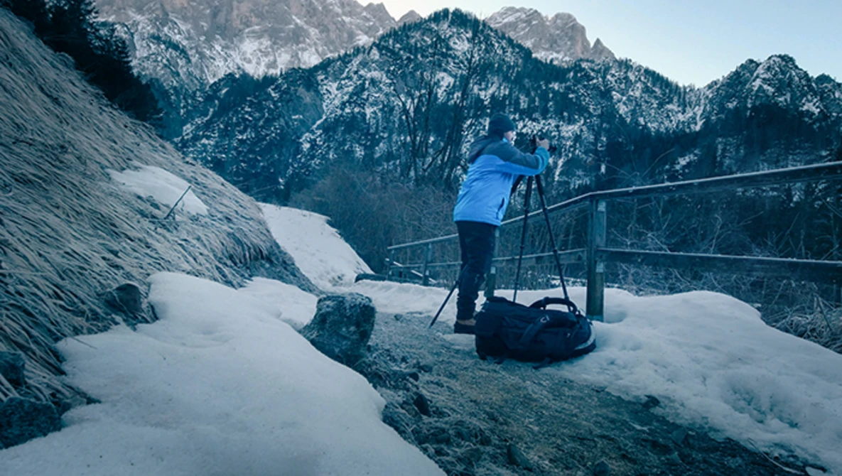 Photographer on a snowy overlook with a tripod and camera, mountains in the distance.