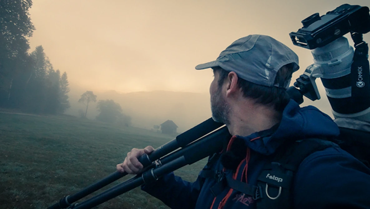 Videographer with camera rig on shoulder filming misty landscape at golden hour.