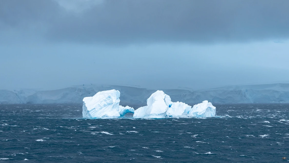 Large blue-white iceberg floating in rough, dark ocean waters with mountains visible in the hazy background.