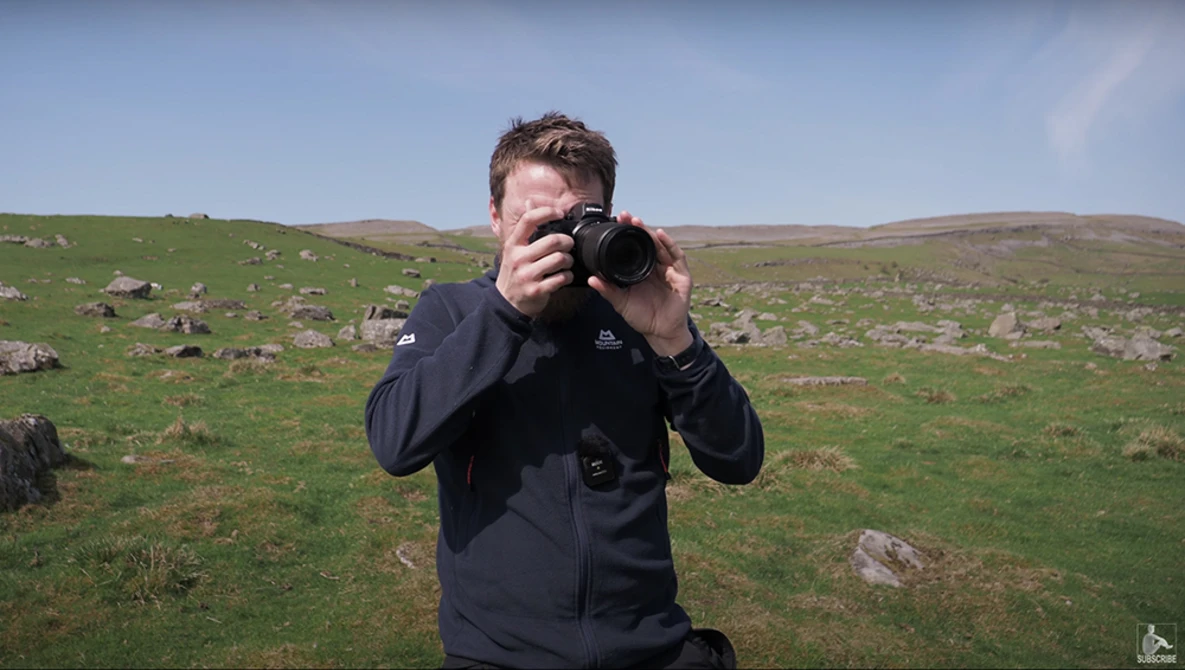 Photographer holding camera to face while standing in open moorland with scattered rocks and green grass.