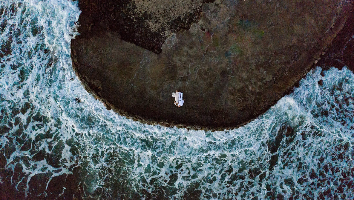 Aerial view of a small white boat in a dark cove surrounded by turquoise ocean waves and rocky coastline.