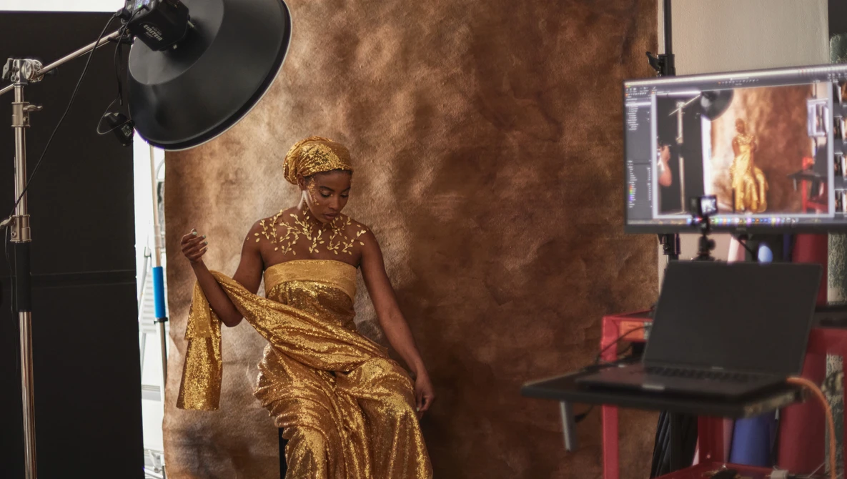 Woman in gold dress and headwrap posing on set with professional studio lighting and textured backdrop.