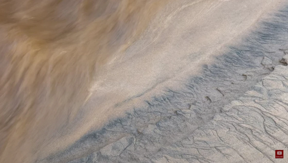 Aerial view of rippled sand patterns and water-carved channels on a desert or beach surface.