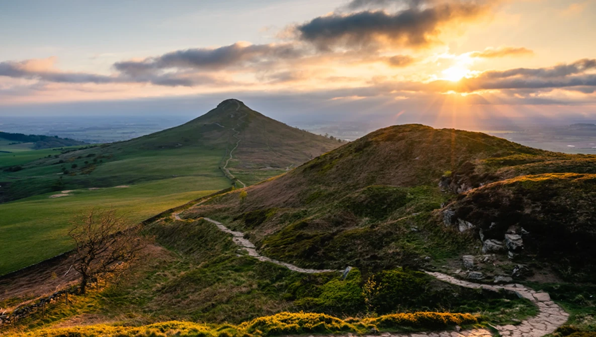 Conical mountain with winding path under golden sunset sky, distant valley landscape below.
