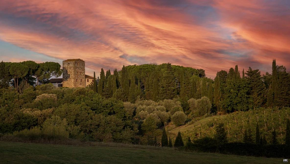 Medieval stone tower on a hillside surrounded by cypress trees and dense vegetation at sunset.