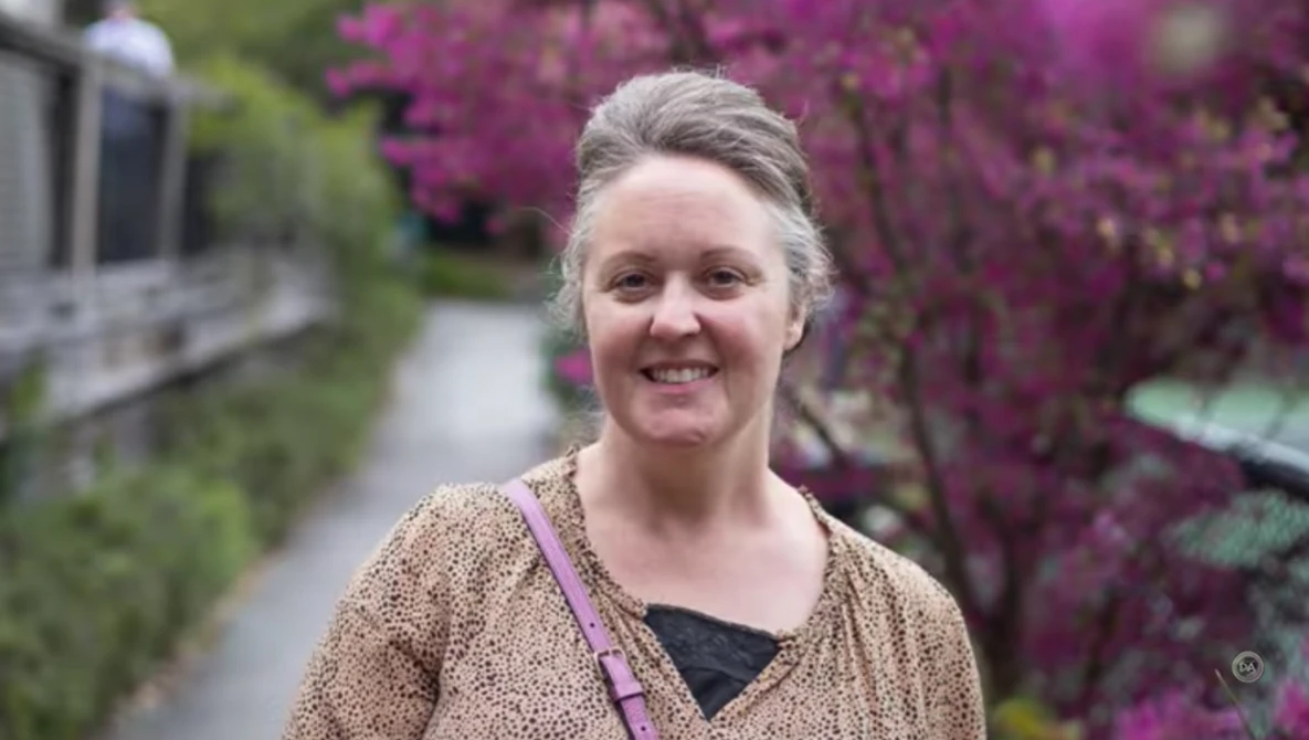 Woman smiling outdoors with vibrant purple flowering shrubs in soft, natural daylight.