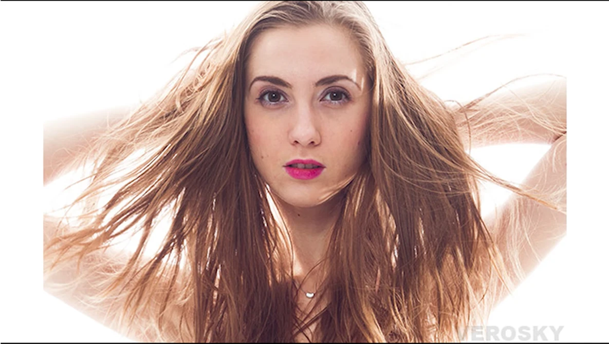 Woman with long brown hair and magenta lipstick, hands raised to head against bright backlit background.