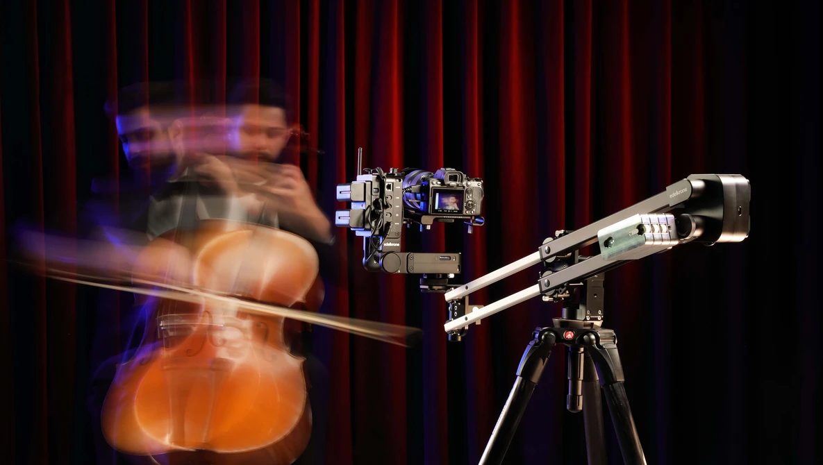 Motion-blurred cellist performing in front of a camera mounted on a tripod against a red curtain backdrop.