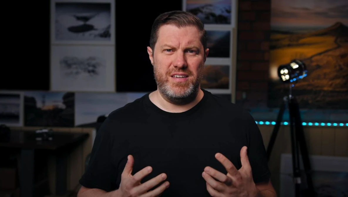Man in black t-shirt speaking to camera in a studio setting with professional lighting equipment visible.
