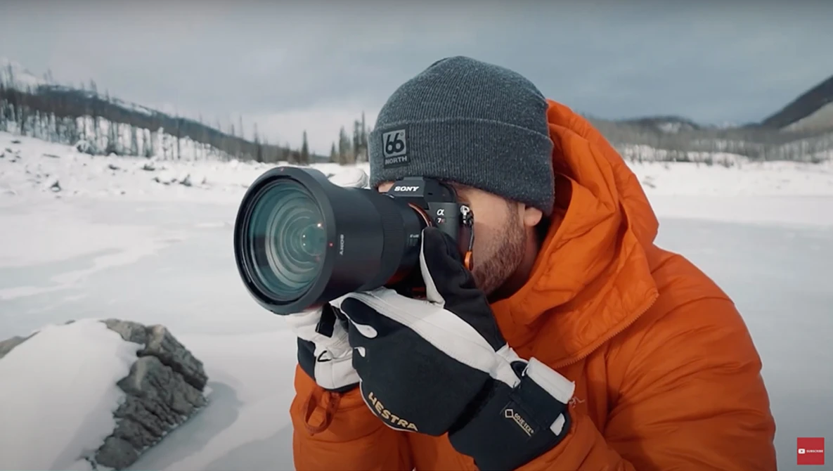 Photographer in orange jacket and gray beanie holding DSLR camera with telephoto lens while shooting on snow-covered landscape.