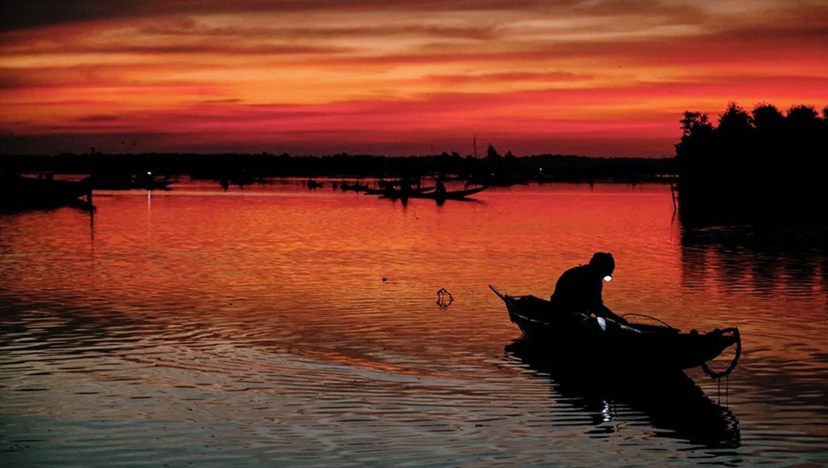 Silhouetted fisherman in small boat on calm water at dramatic sunset with fiery orange and red sky.