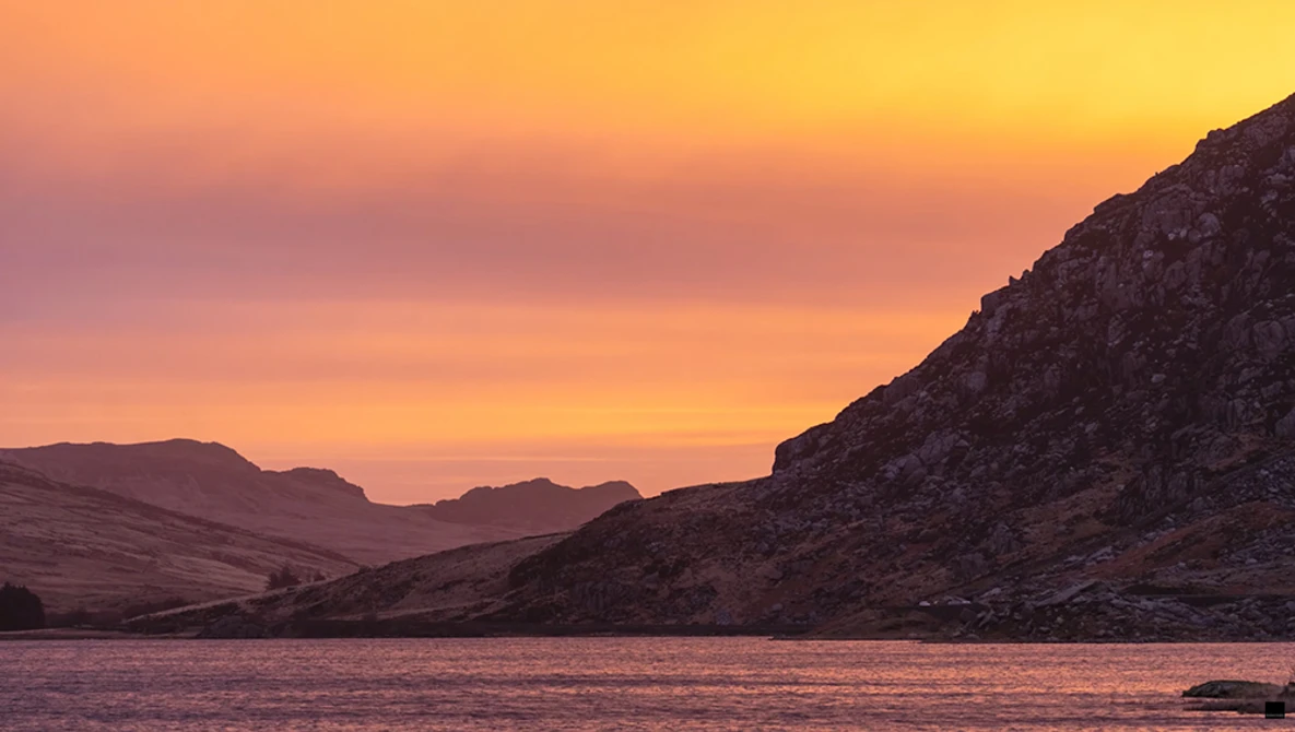 Rocky mountainous shoreline at sunset with warm golden and pink sky reflected in still water.