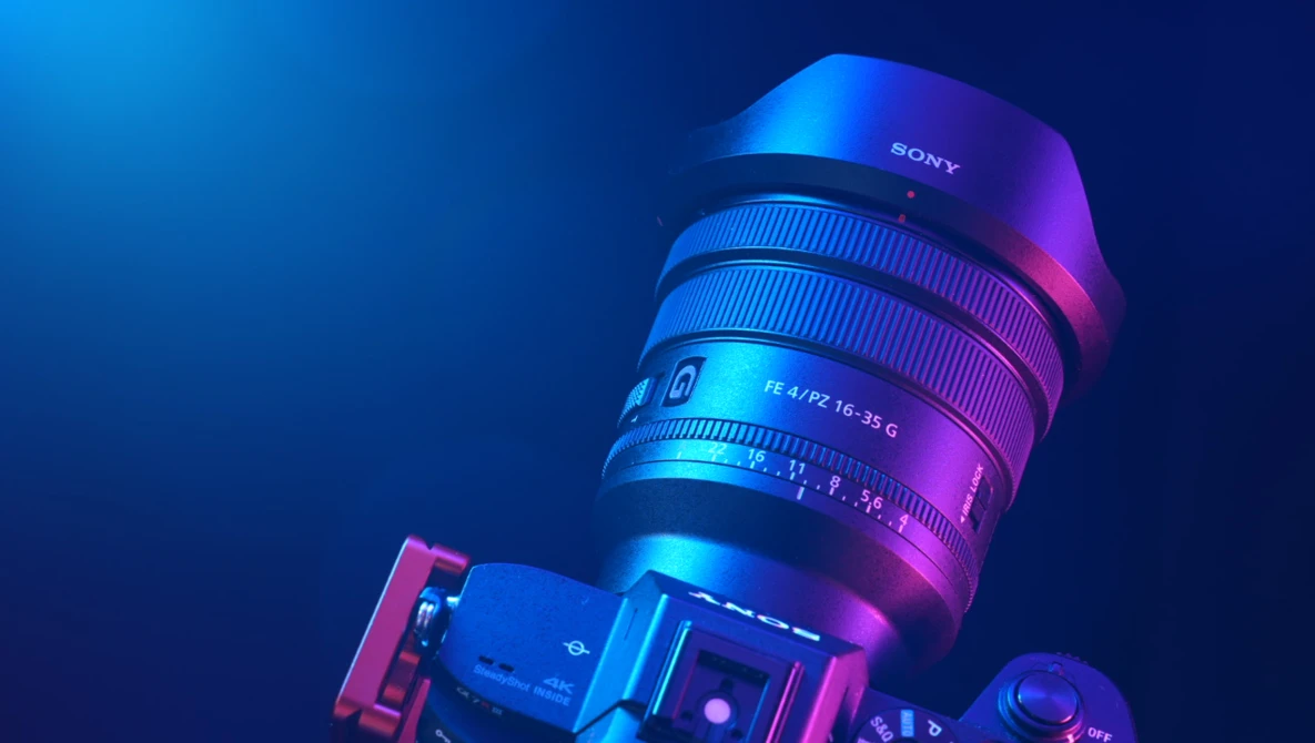 Sony camera lens mounted on camera body, illuminated with blue and magenta neon lighting against dark background.