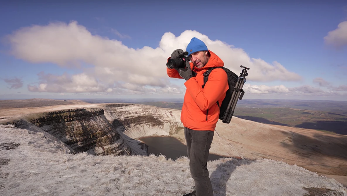 Photographer in orange jacket and blue cap holding camera, standing on white chalk cliff overlooking vast landscape.