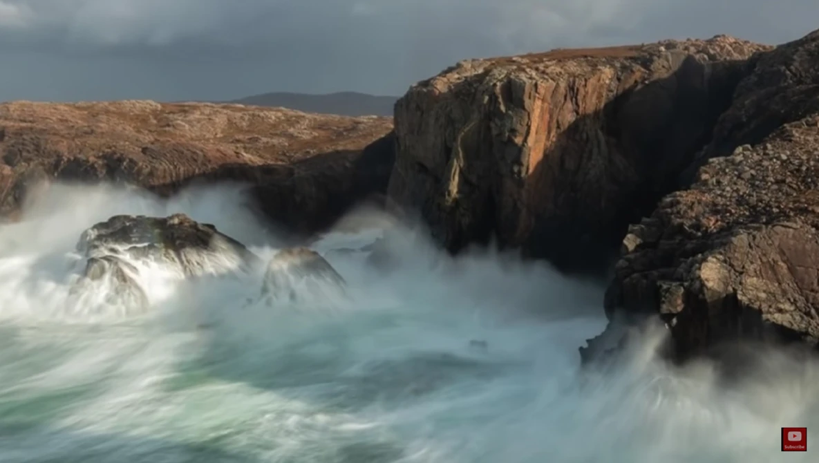 Turbulent ocean waves crash against rocky cliffs under overcast sky.