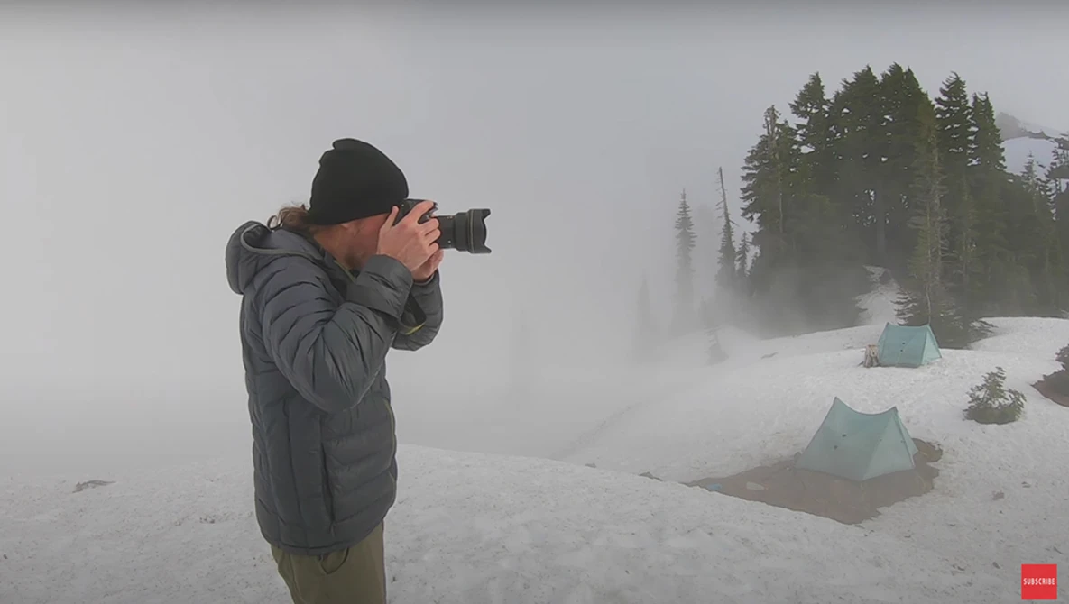 Photographer in dark winter jacket and black beanie capturing images in heavy fog and snow with distant tent visible.