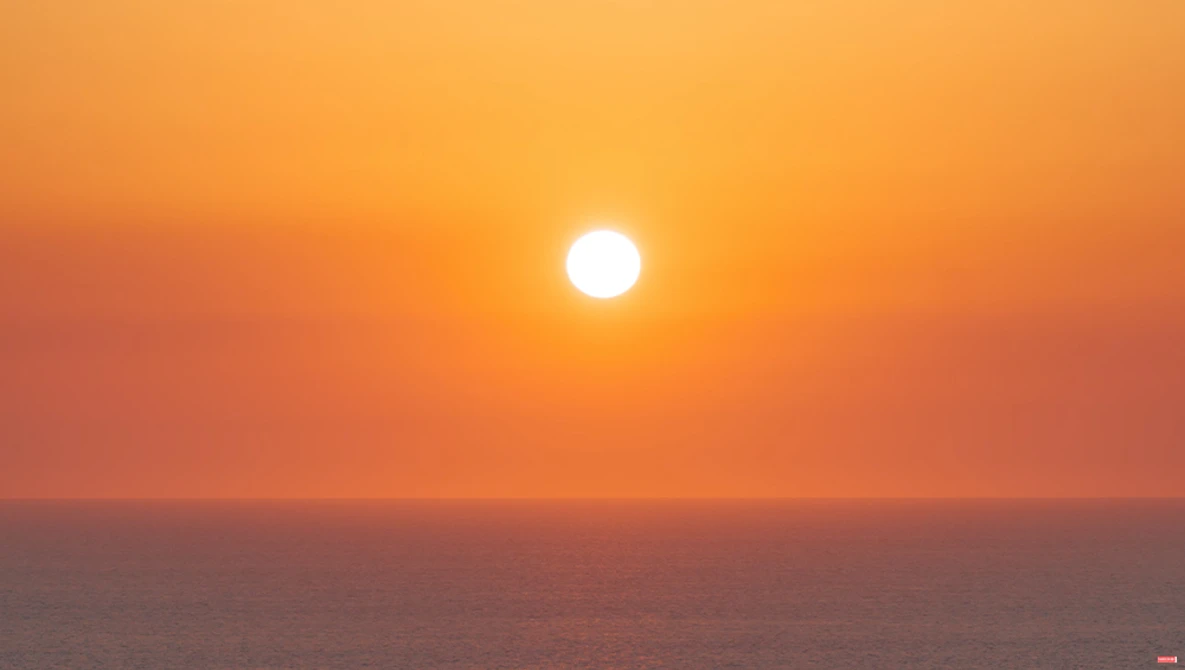 Bright sun descending toward the ocean horizon under a gradient orange and pink sky at sunset.
