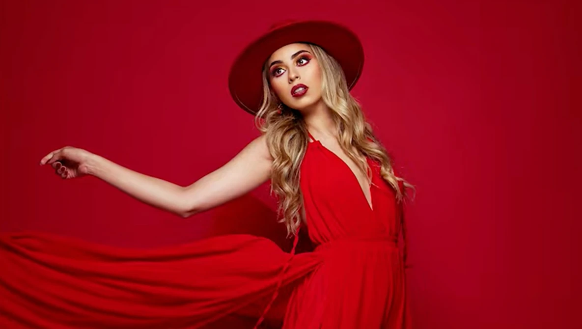 Woman in red dress and burgundy hat against solid red background, arm extended gracefully.