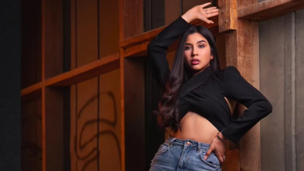 Woman in black crop top and jeans posed against wooden architectural elements with warm ambient lighting.