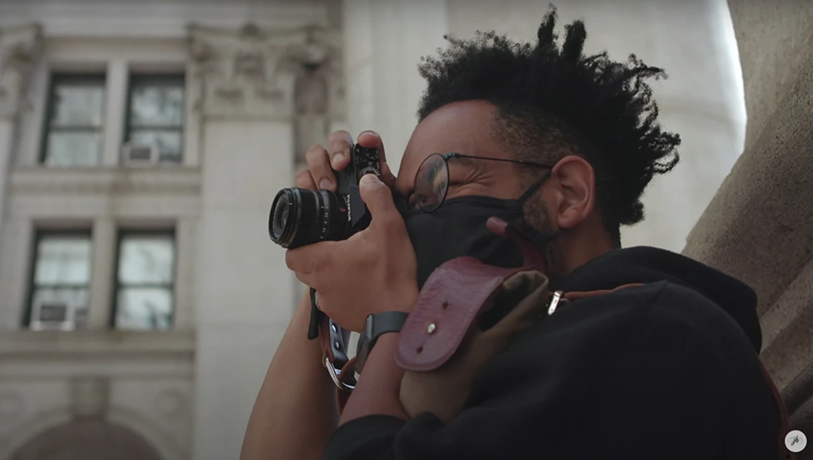 Photographer wearing glasses and a pink camera strap looks through a mirrorless camera viewfinder while positioned against a building facade.