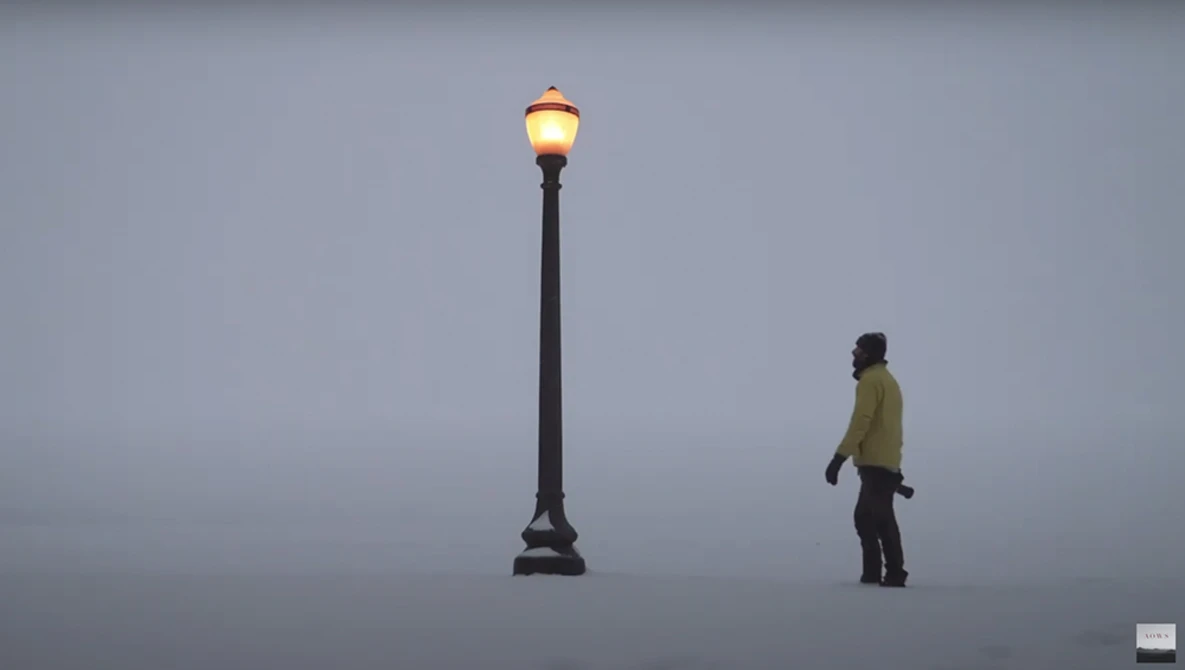 Person in yellow jacket standing near an illuminated street lamp against a gray sky.