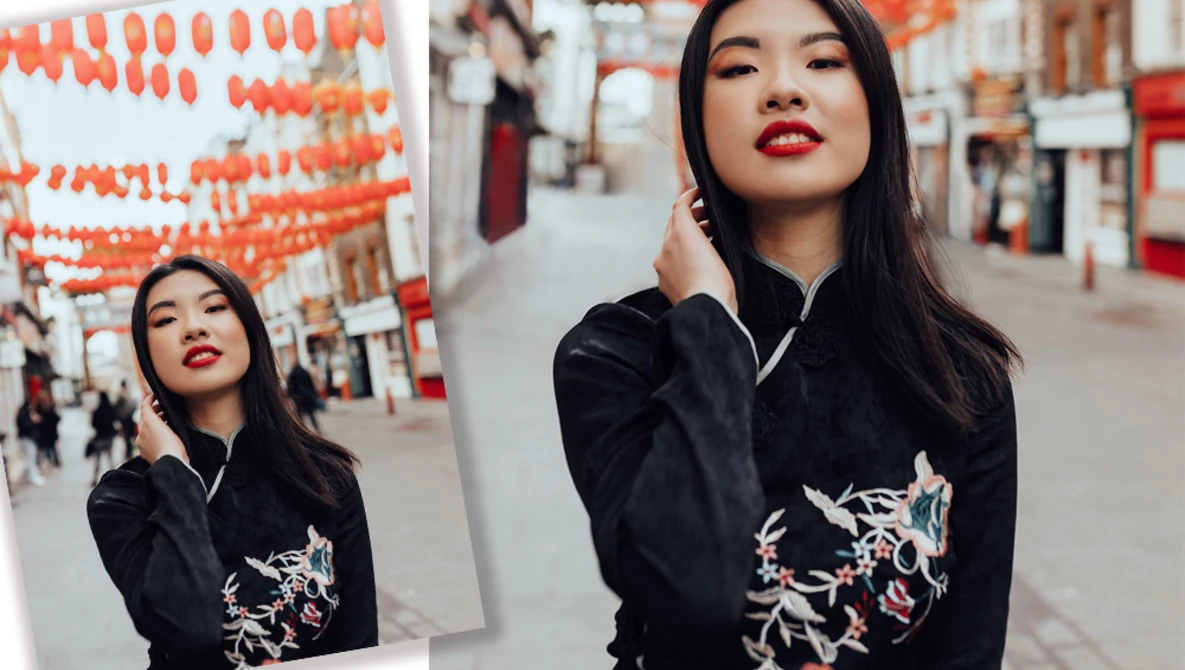 Portrait of a woman with long dark hair and red lipstick wearing a black jacket with embroidered dragons, shot against a street backdrop with orange lanterns.