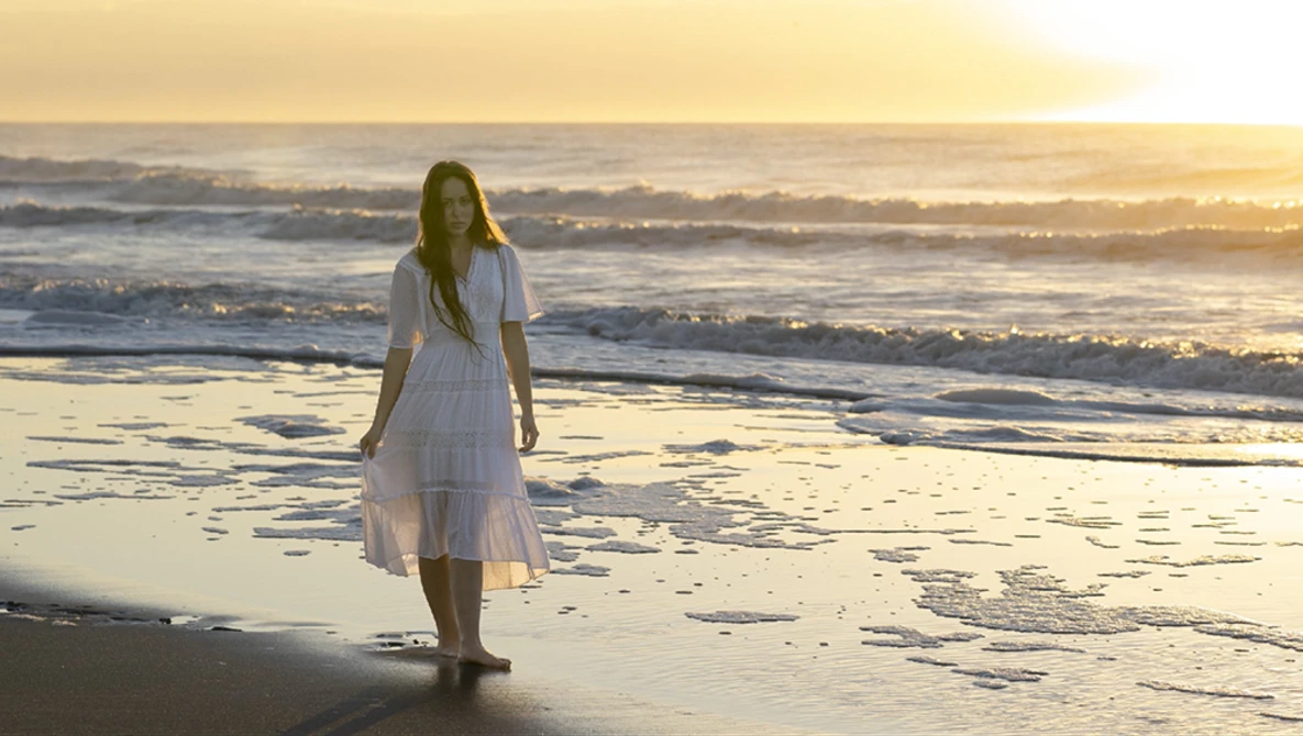 Woman in white dress standing in shallow ocean water during golden hour sunset.