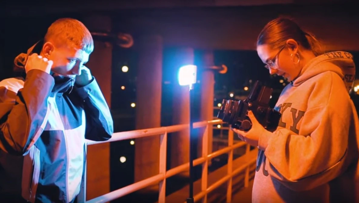 Two people on a metal railing at night, one singing into a microphone while the other operates a camera.