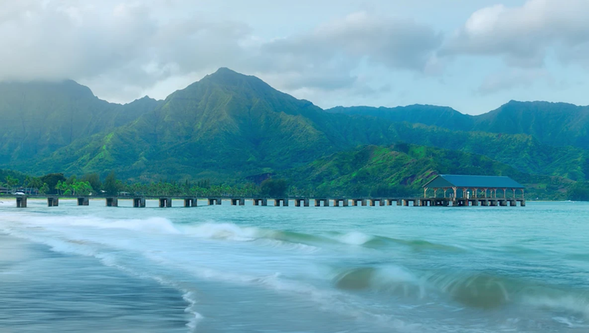Wooden pier extending into turquoise ocean waters with green mountains rising dramatically behind it under partly cloudy skies.