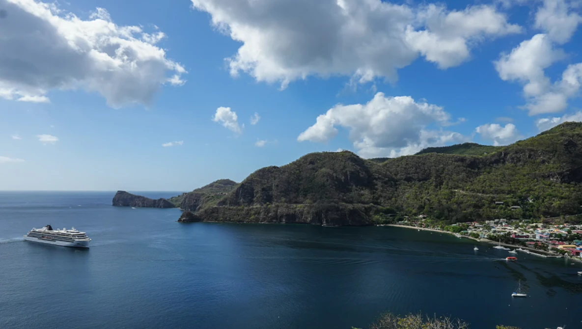 Tropical coastal bay with lush green mountainside, cruise ship anchored offshore, and small waterfront settlement nestled along the shoreline.
