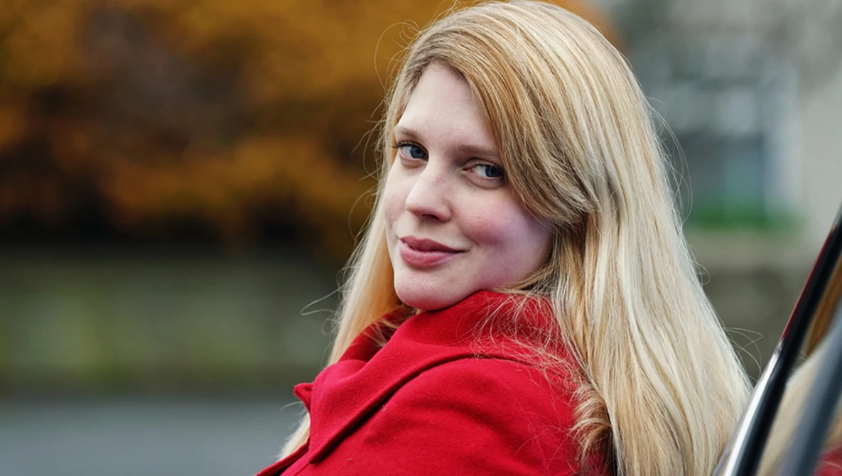 Woman with blonde hair wearing a red jacket, looking over her shoulder at the camera with a slight smile.