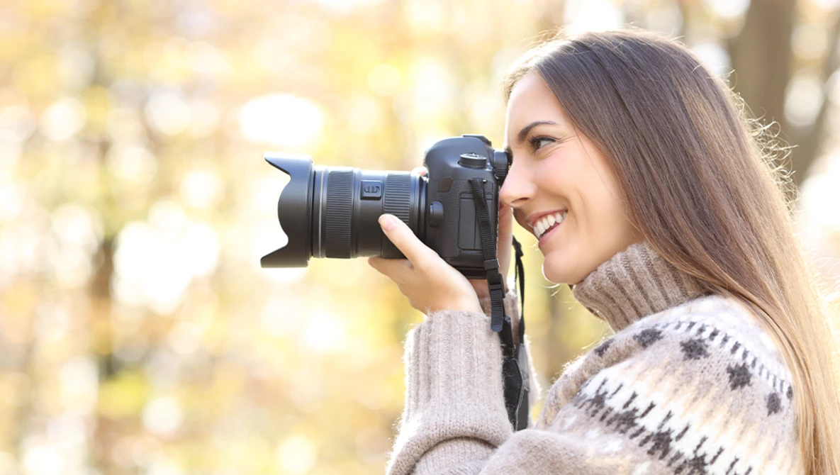 Female photographer holding a mirrorless camera with telephoto lens to her eye, smiling while shooting outdoors.