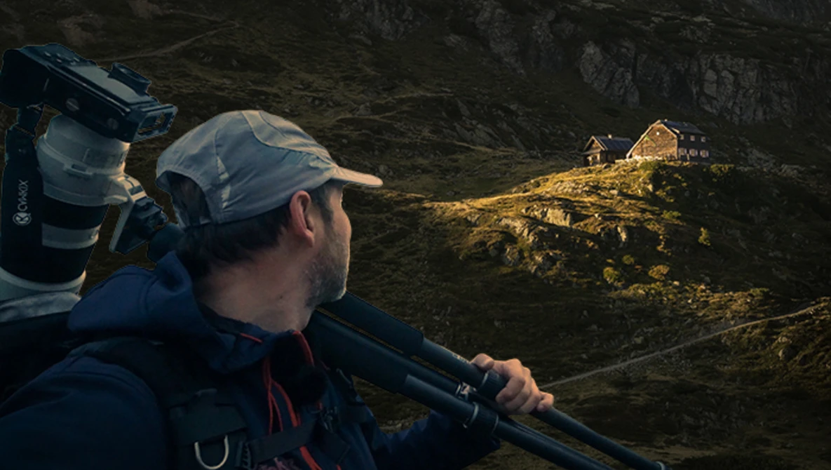 Photographer with telephoto lens aimed at illuminated buildings on a distant mountainside at dusk.