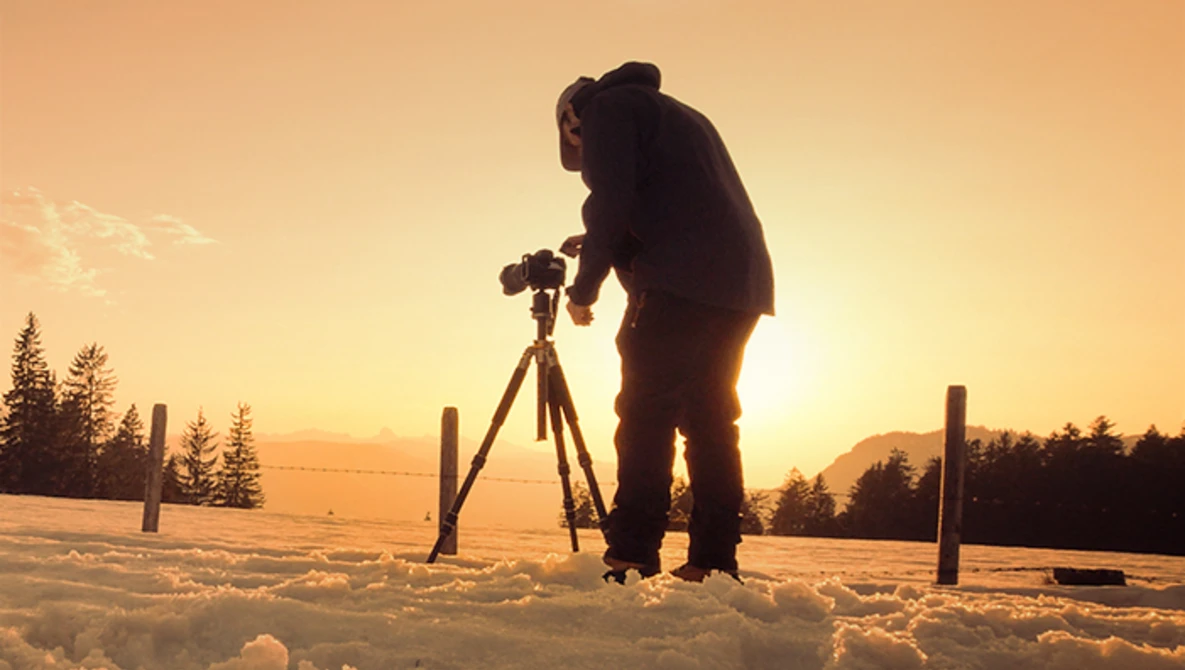 Silhouetted photographer operating a camera on tripod in snowy landscape at golden hour.