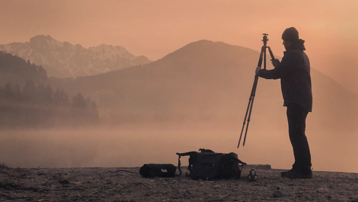 Silhouetted surveyor operating a theodolite on a tripod at sunrise, with misty mountains and forested terrain in the background.