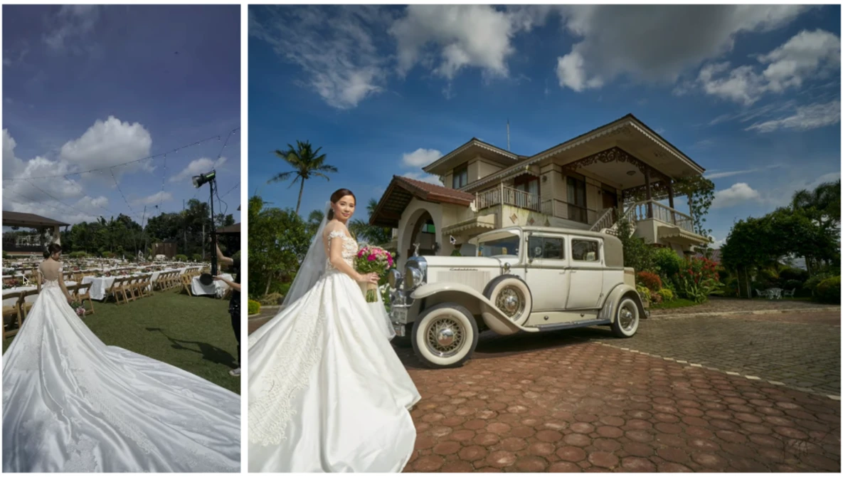 Bride in flowing white gown posing beside vintage cream-colored car in front of colonial mansion.