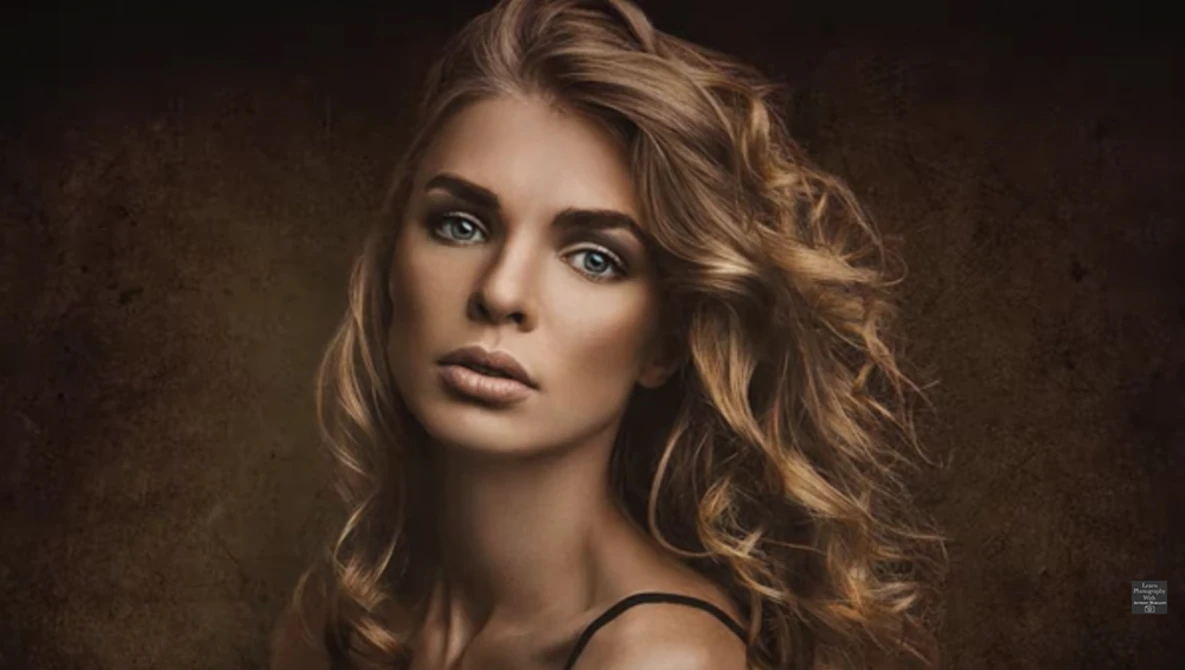 Portrait of a woman with wavy blonde hair against a textured brown background.