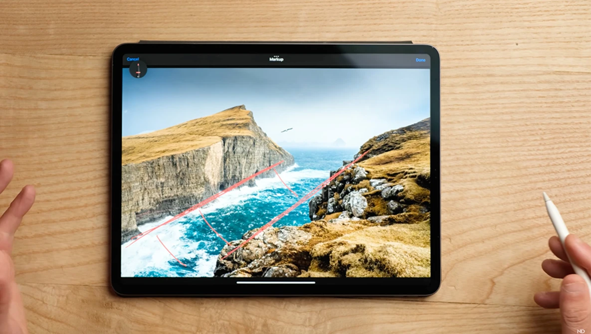 Tablet displaying a coastal landscape with dramatic cliffs and turquoise water, held by hands on a wooden surface.