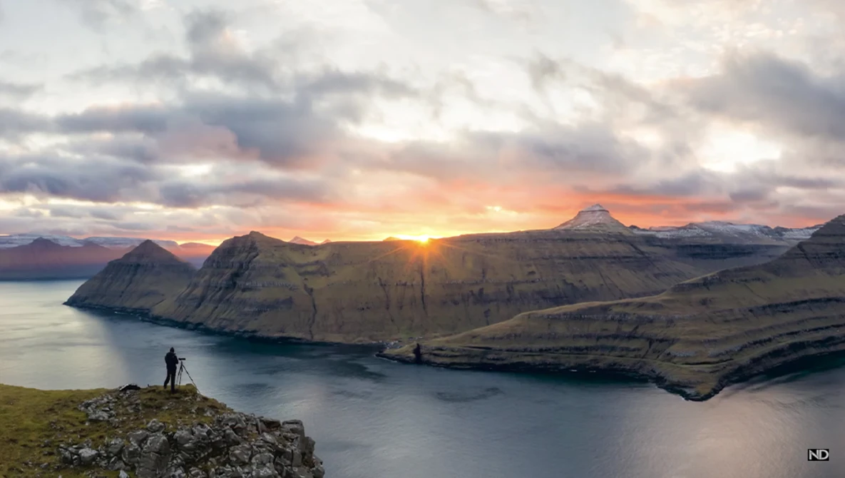 Photographer standing on rocky outcrop overlooking fjord at sunset with snow-capped mountains.