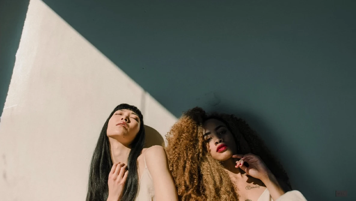 Two women with long hair looking upward against a teal wall with dramatic window light.