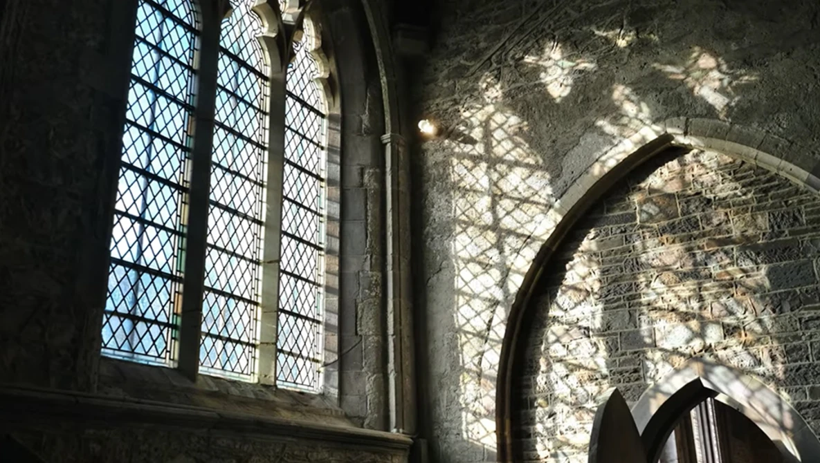 Gothic stone interior with arched doorway and latticed window casting dramatic shadows on textured walls.