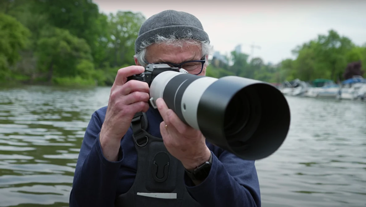 Photographer holding a camera with a large telephoto lens while standing in a boat on the water.