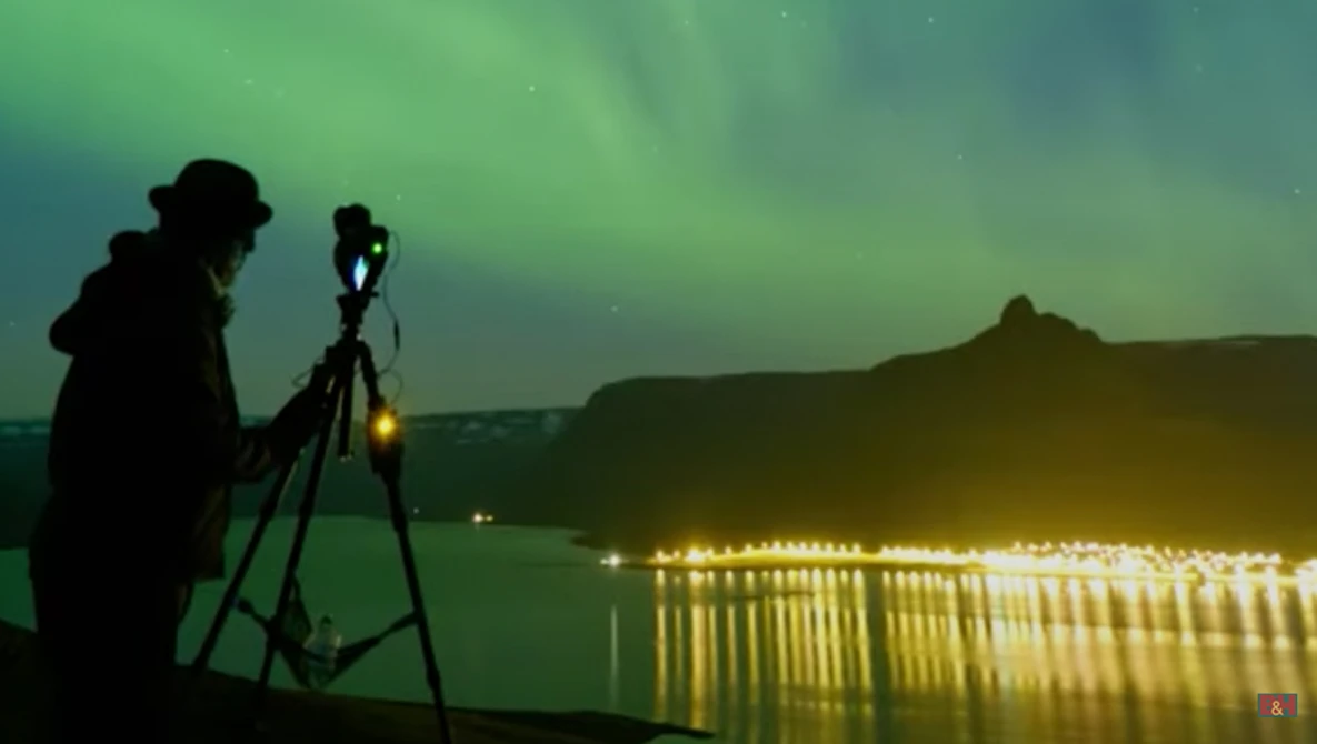 Photographer on tripod capturing northern lights over illuminated lakeside landscape at night.