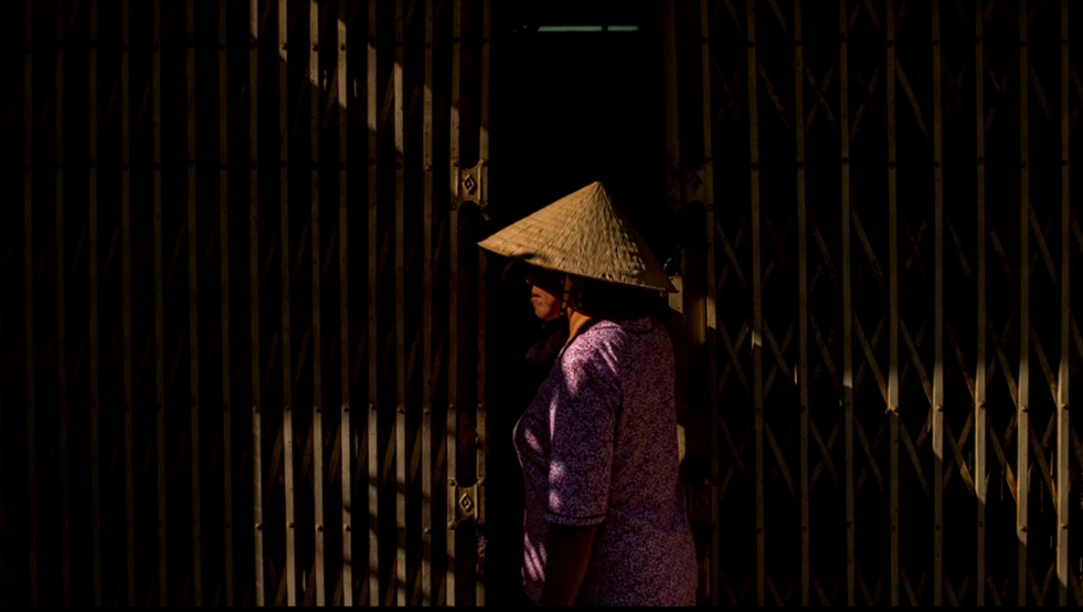 Person wearing traditional cone hat and purple garment standing in dramatically lit narrow corridor with wooden walls.