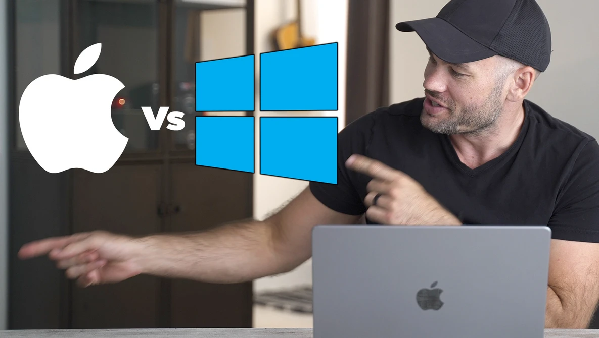 Man in black cap pointing at laptop with Apple vs Windows logos displayed on screen.