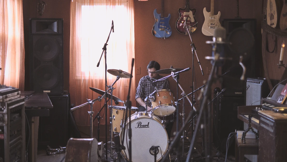 Drummer performing in a home studio surrounded by guitars and musical equipment with warm window lighting.
