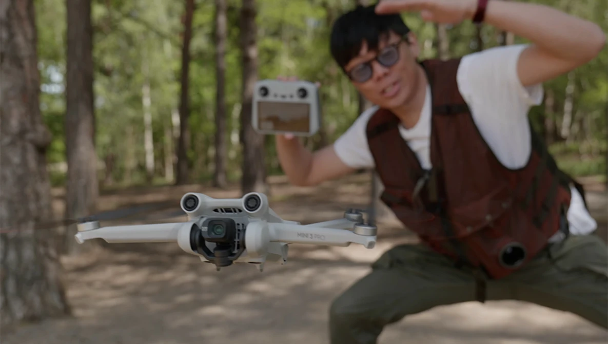 Photographer holding DJI Mini drone controller while operating a white quadcopter in a wooded area.