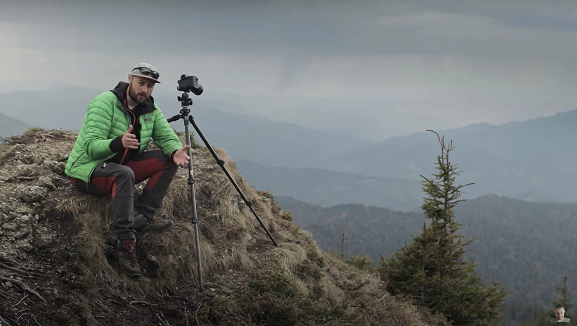 Photographer crouched on a rocky ridge with camera on tripod, overlooking layered mountain ranges in mist.