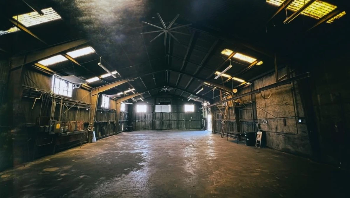 Large abandoned warehouse interior with industrial ceiling, metal scaffolding, and scattered windows casting warm light across concrete floor.